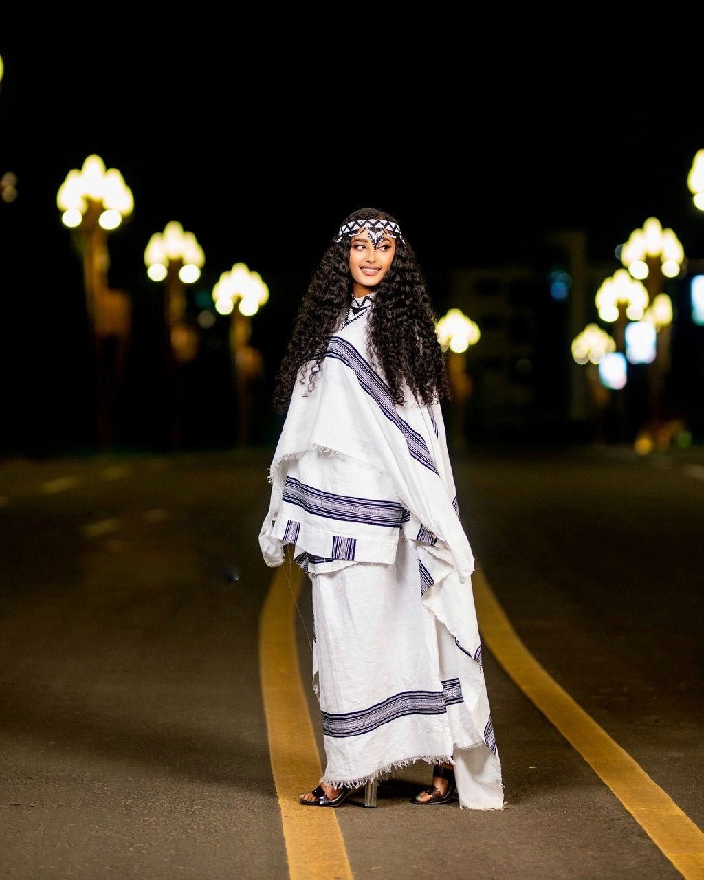 Woman in a white and blue traditional outfit standing on a road at night with lights in the background