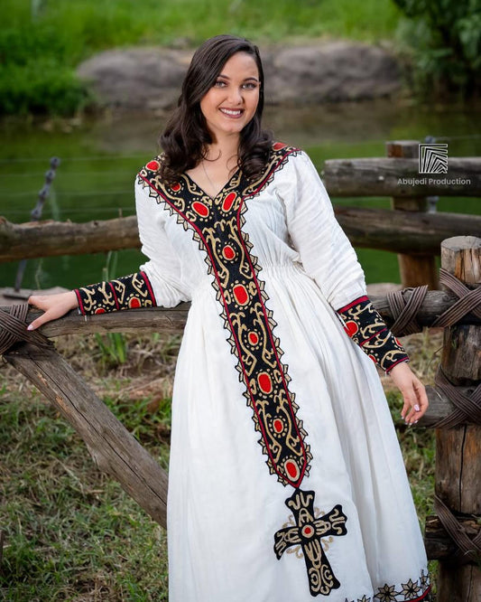 Woman wearing a white dress with intricate black and red embroidery, standing outdoors by a wooden fence.