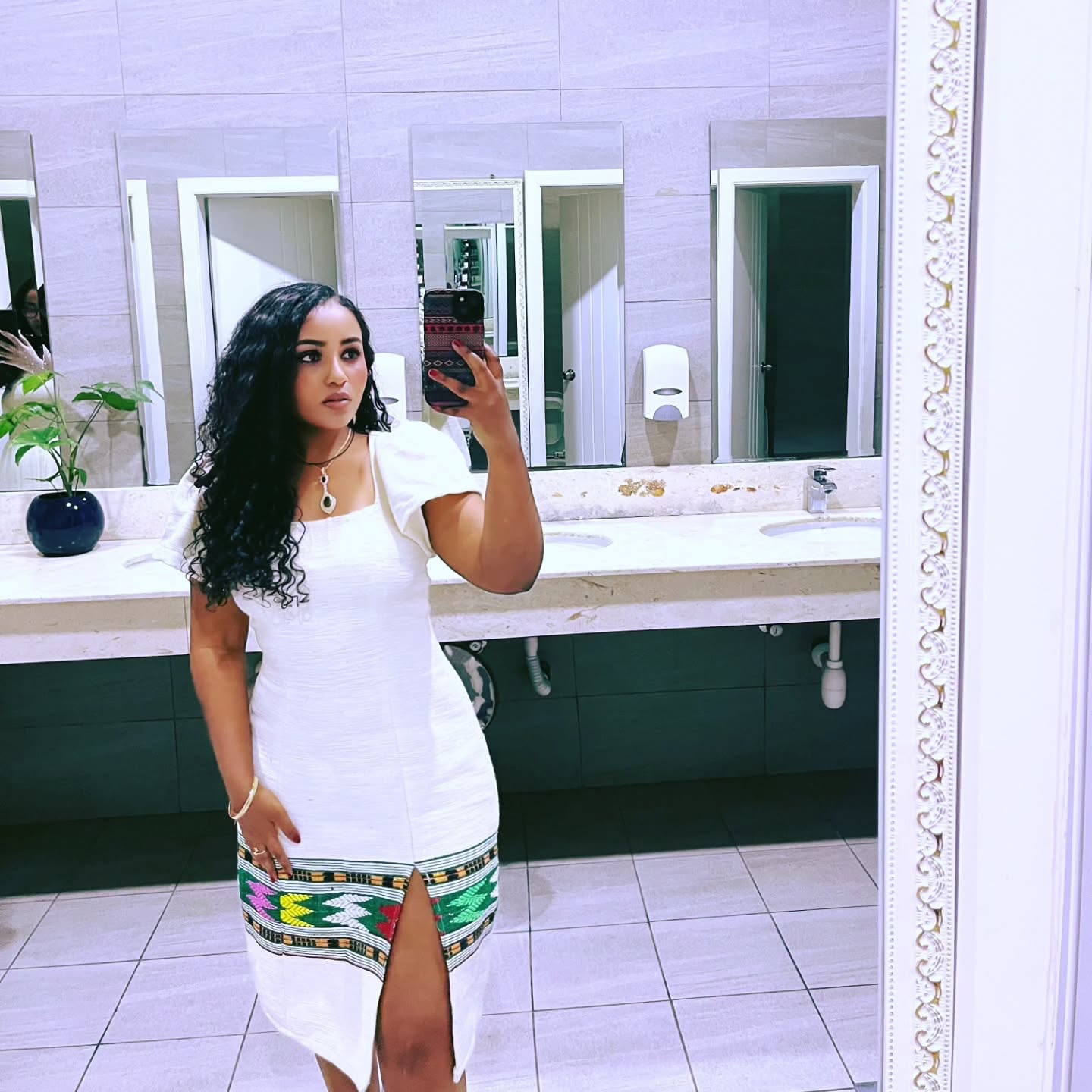 Woman taking a mirror selfie in a bathroom wearing a white dress with a colorful hem.