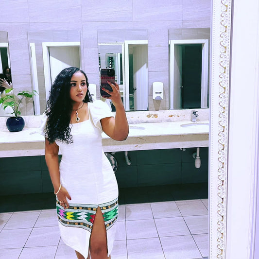 Woman taking a mirror selfie in a bathroom wearing a white dress with a colorful hem.