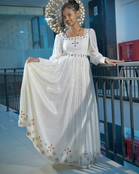 Woman in a long white dress with floral embroidery standing on a balcony.