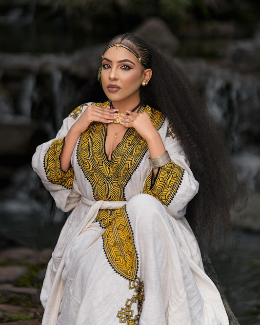 Woman wearing a white and gold traditional outfit with jewelry, standing against a natural background.