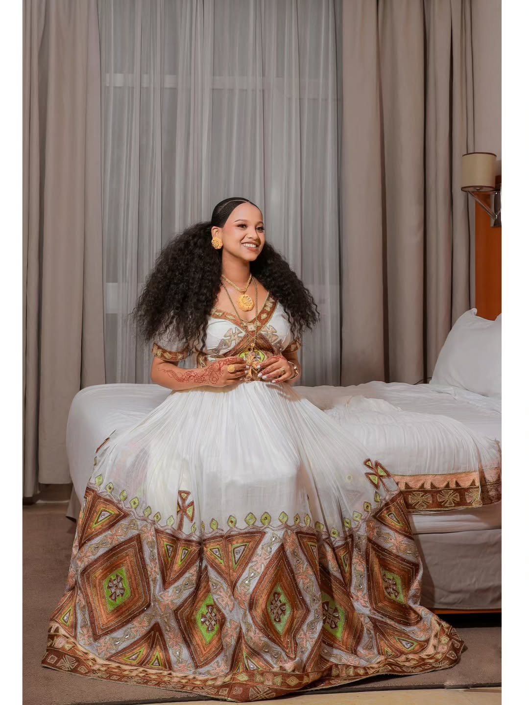 Woman in a white and patterned dress sitting on a couch with curtains in the background