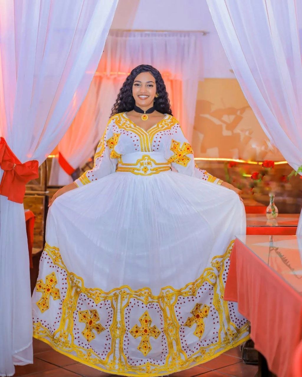 Woman in a white and yellow traditional outfit sitting in a decorated room.