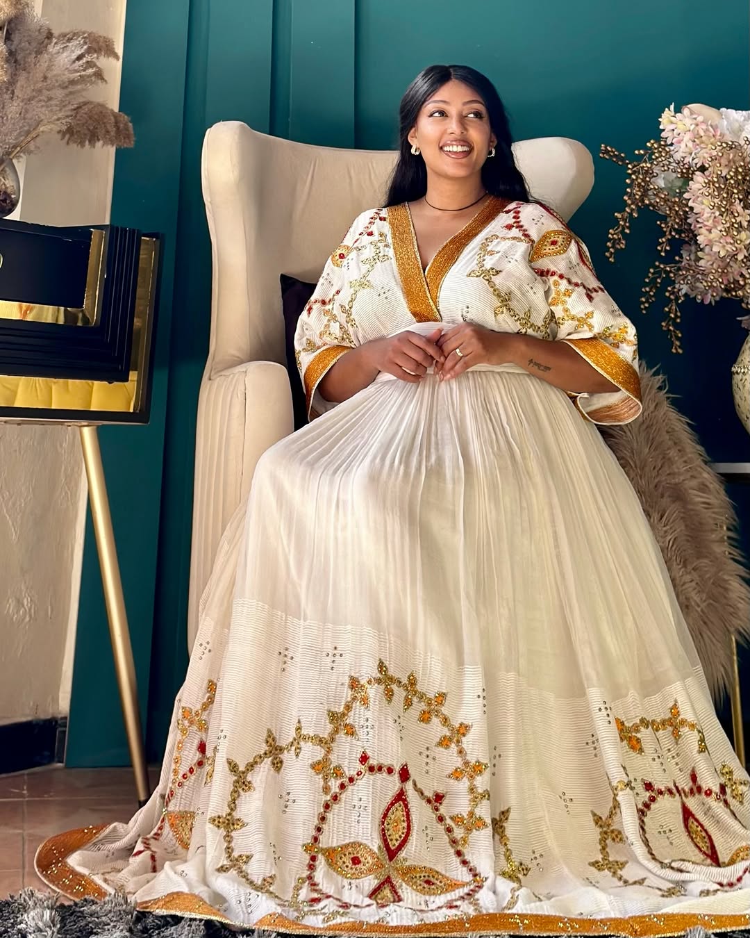 Woman in a white and gold embroidered saree sitting on a chair against a teal wall.
