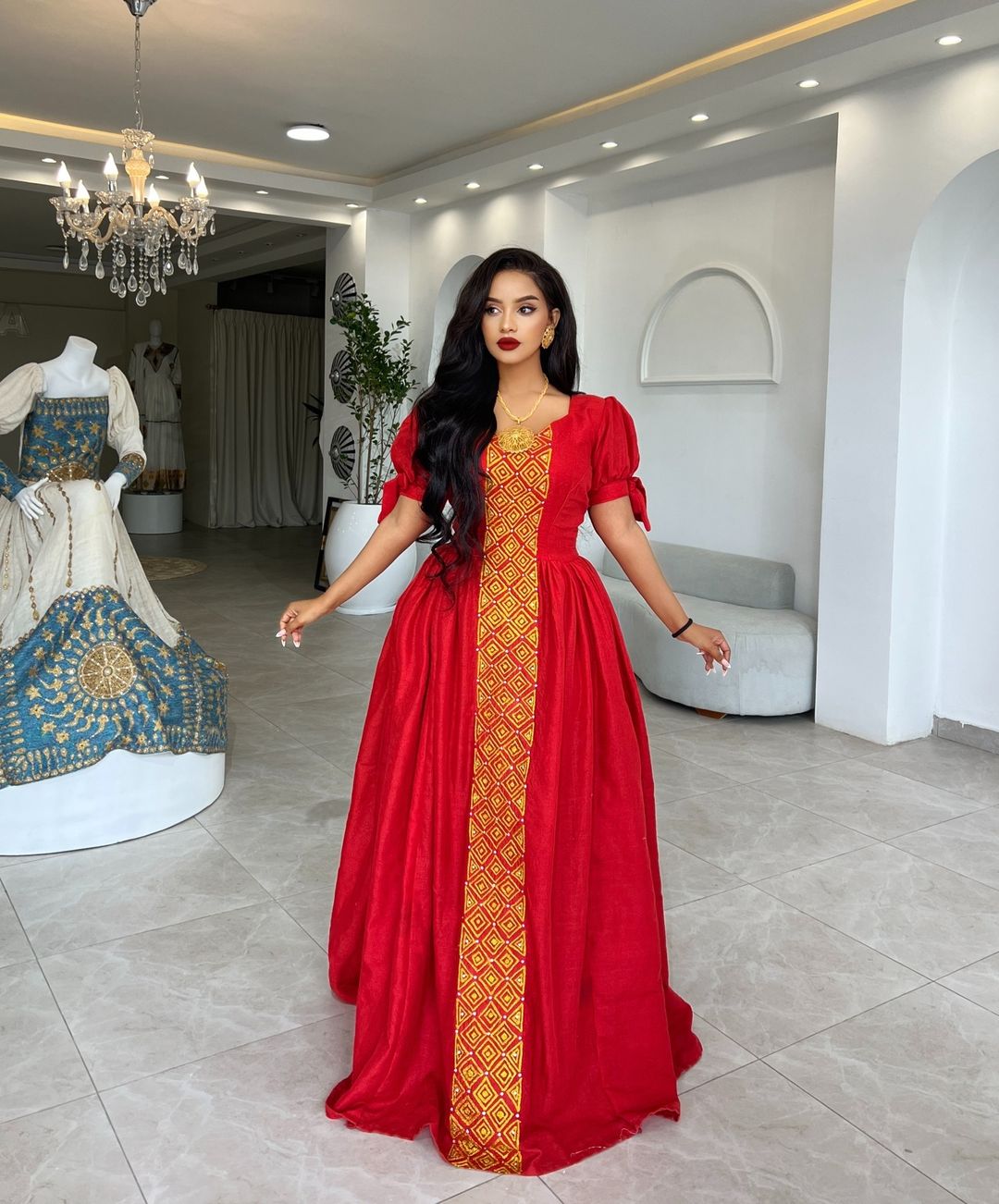 Woman in a red dress with gold patterns standing in a stylish interior setting.