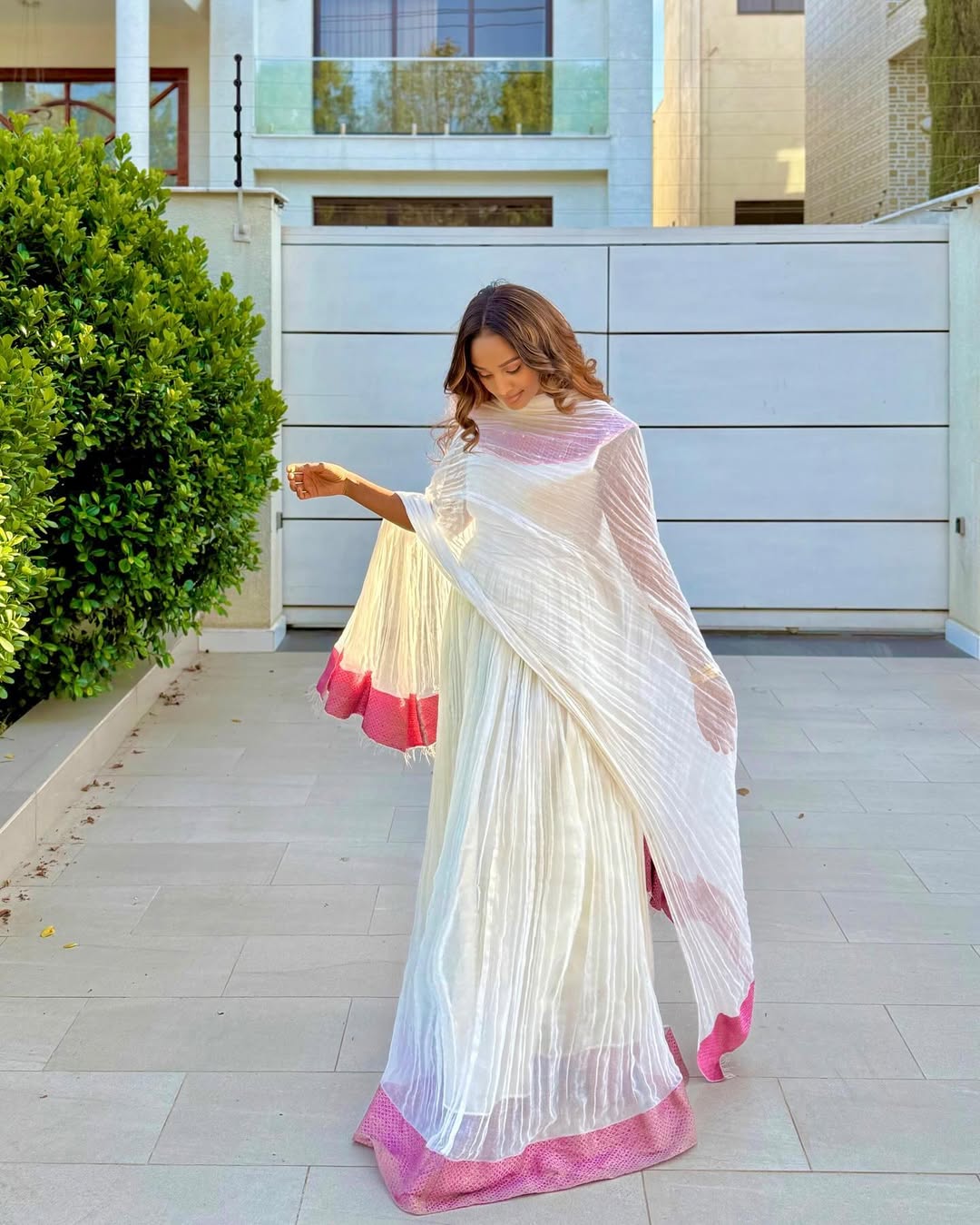Woman in a white and pink saree standing outside a building.