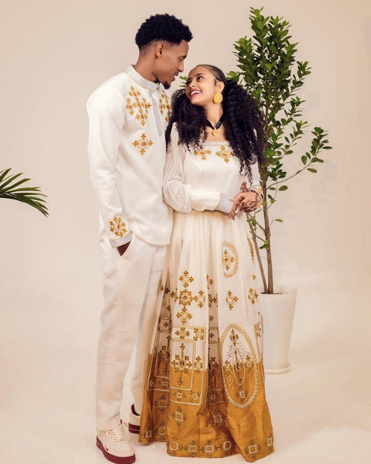 Couple in matching white outfits with gold patterns standing next to a plant.