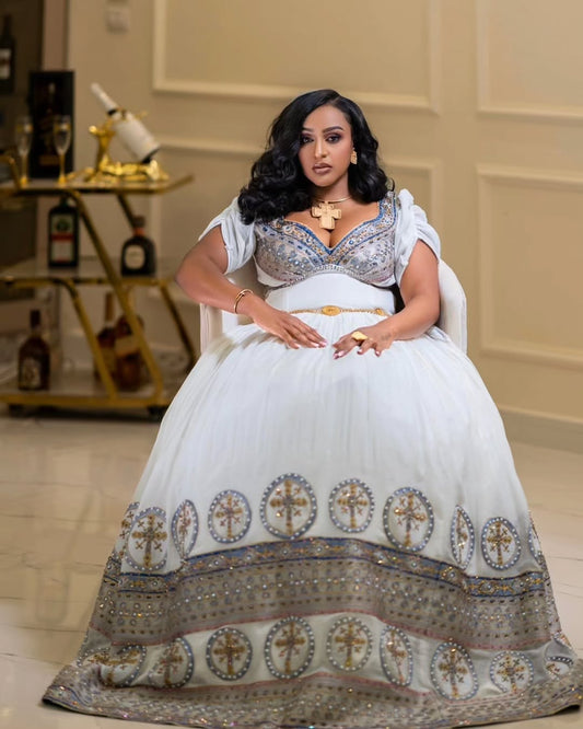 Woman in a white and gold traditional outfit sitting in a room with decor elements.