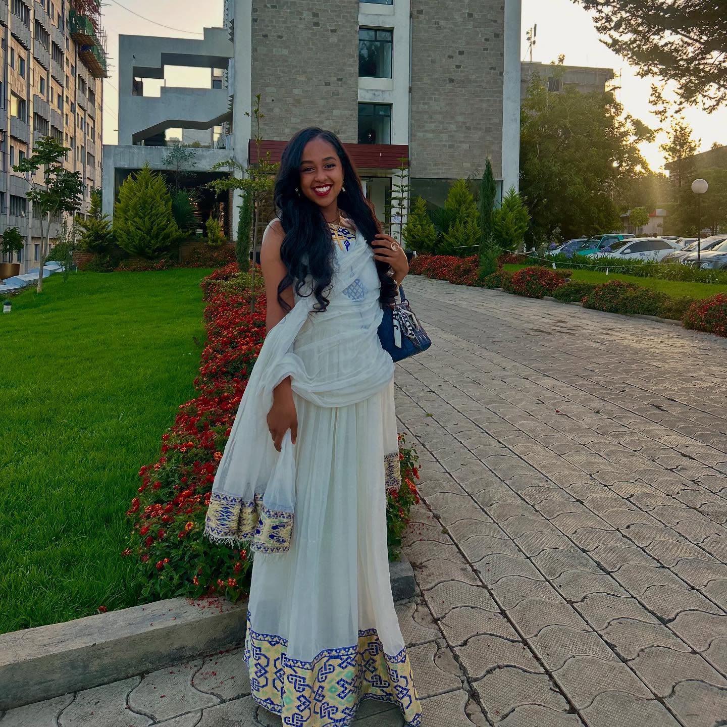 Woman in a white and blue traditional outfit standing on a pathway with greenery and buildings in the background.