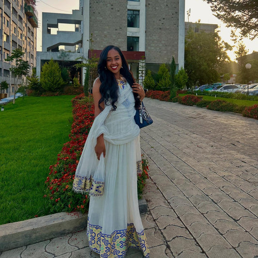 Woman in a white and blue traditional outfit standing on a pathway with greenery and buildings in the background.