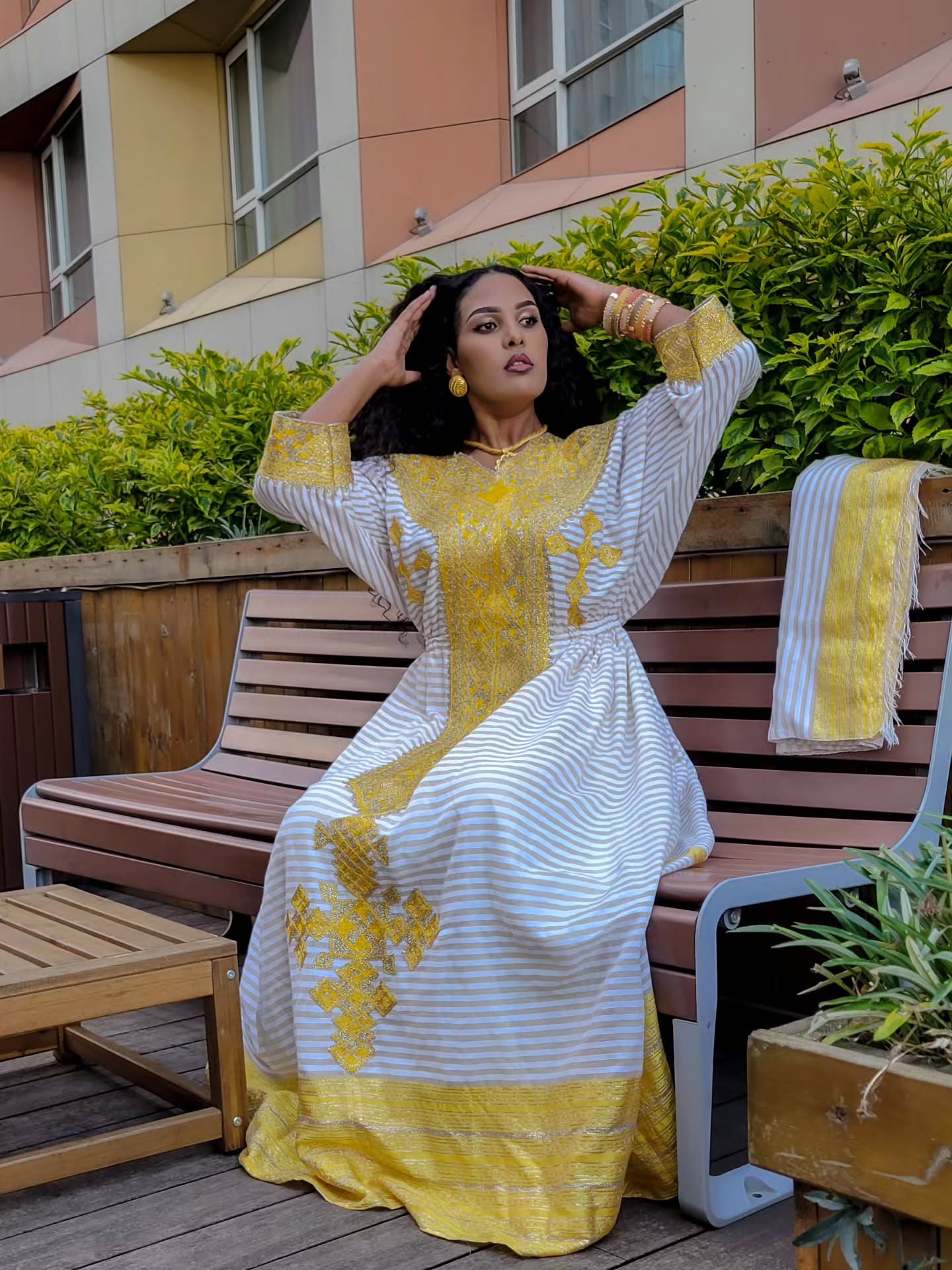 Woman in a yellow and white dress sitting on a bench with greenery and a building in the background