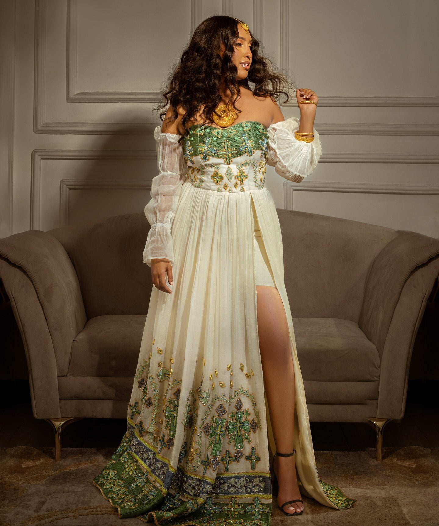 Woman in a long, elegant dress with floral patterns in a room with a gray sofa.