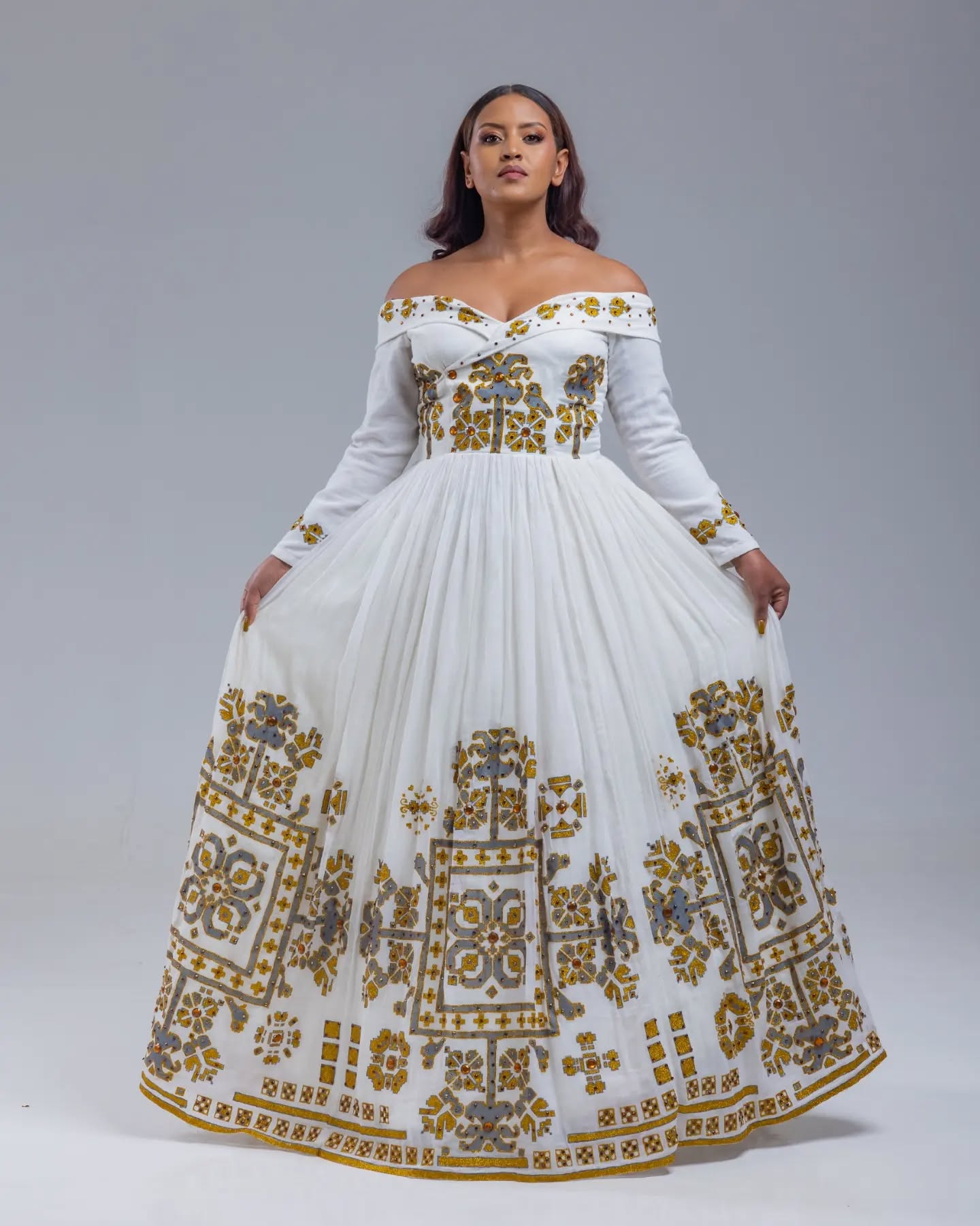 Woman wearing a white off-shoulder dress with gold embroidery on a gray background