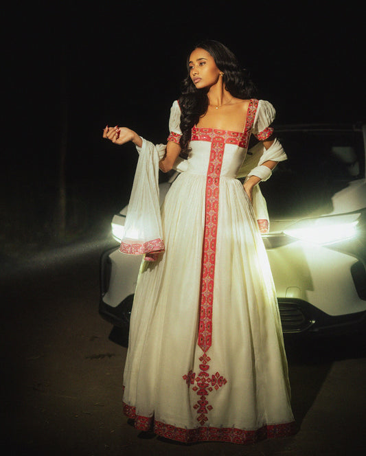Woman in a white dress with red embroidery standing in front of a car at night.
