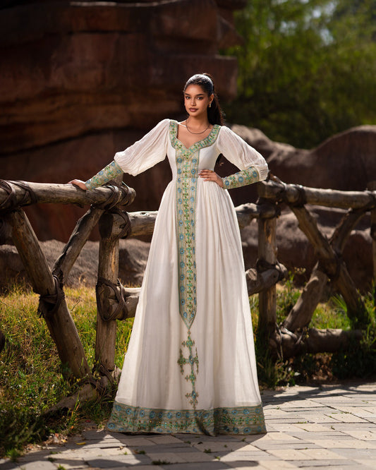 Woman in a white dress with green patterns standing outdoors near wooden fences and trees.