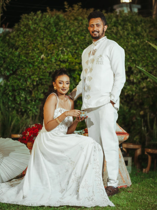 Man and woman in formal white attire sitting outdoors with greenery in the background